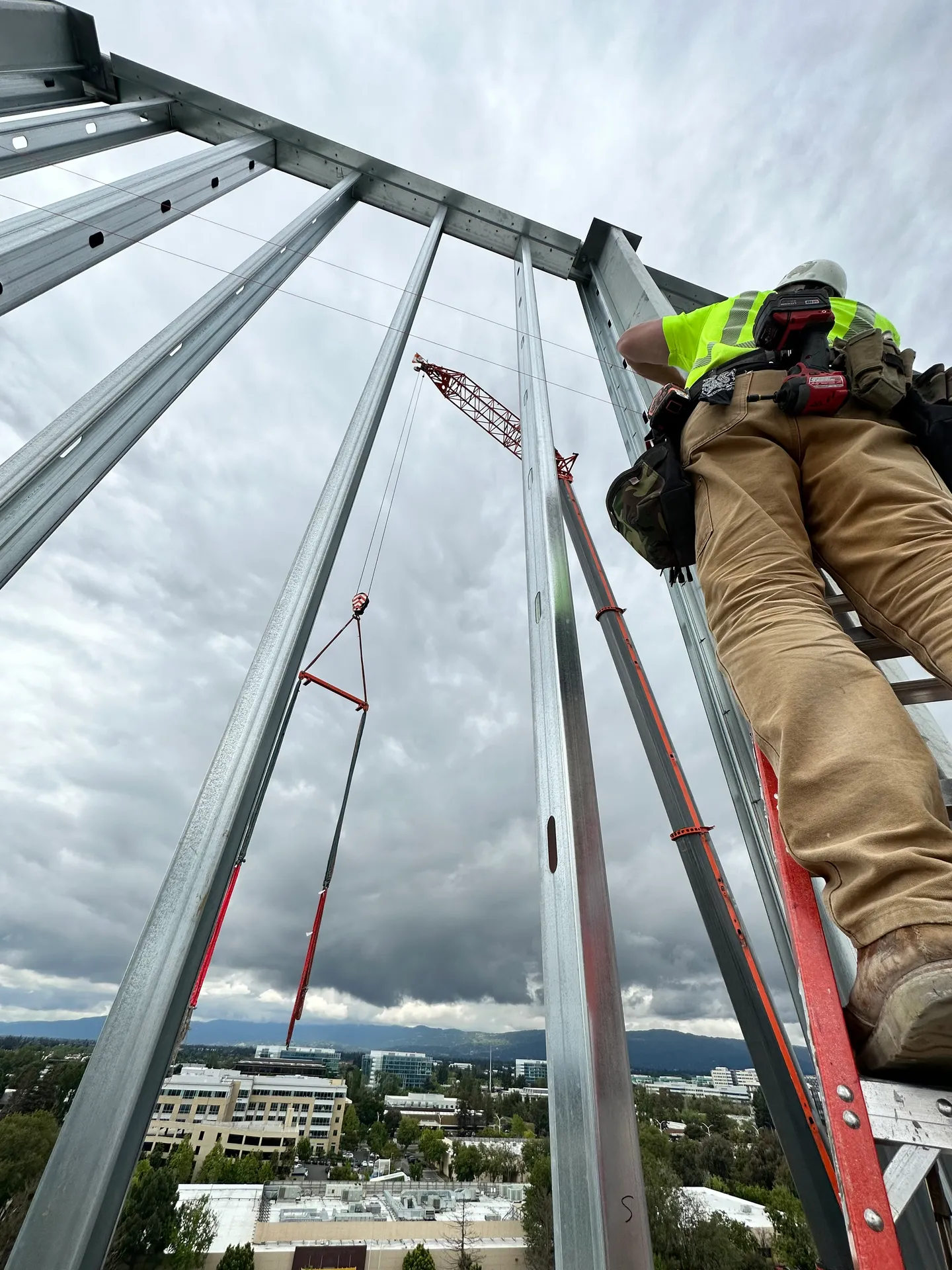 A 93 Electric electrician in a hard hat and hi-vis vest climbs a ladder up steel structural framing high on a commercial high-rise, with a crane lifting a load behind him.